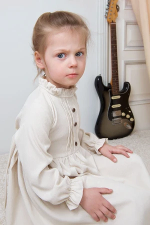 Young girl in a vintage-style cream dress with ruffled collar sitting indoors next to an electric guitar.