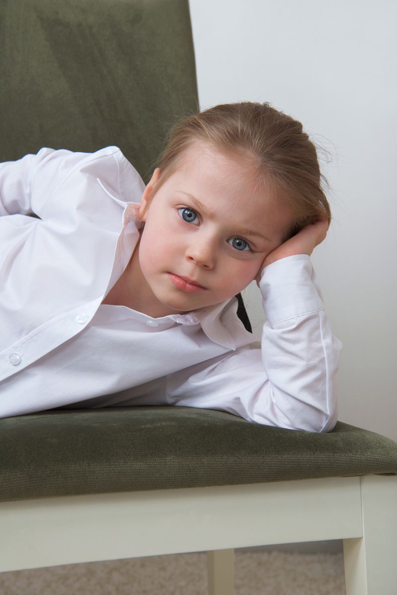 Young child in an oversized white collared shirt resting their chin on their hand while leaning on an olive green chair.