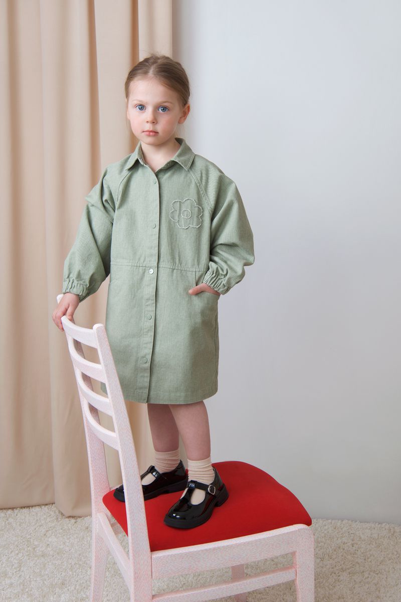 A young girl in a sage green button-up smock dress stands atop a chair with a red seat against a neutral studio backdrop.