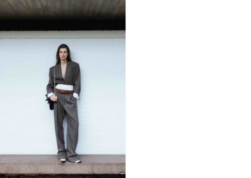 Model Luca Gajdus stands on a stone ledge wearing an oversized grey tailored suit with a white waistband against a white wall.