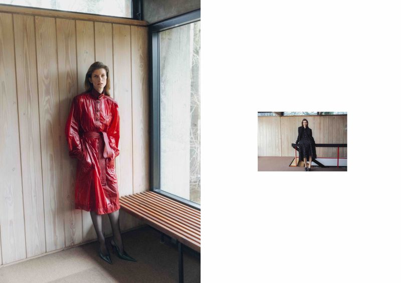 Model Luca Gajdus wearing a belted glossy red trench coat standing beside a slatted wood bench and window.