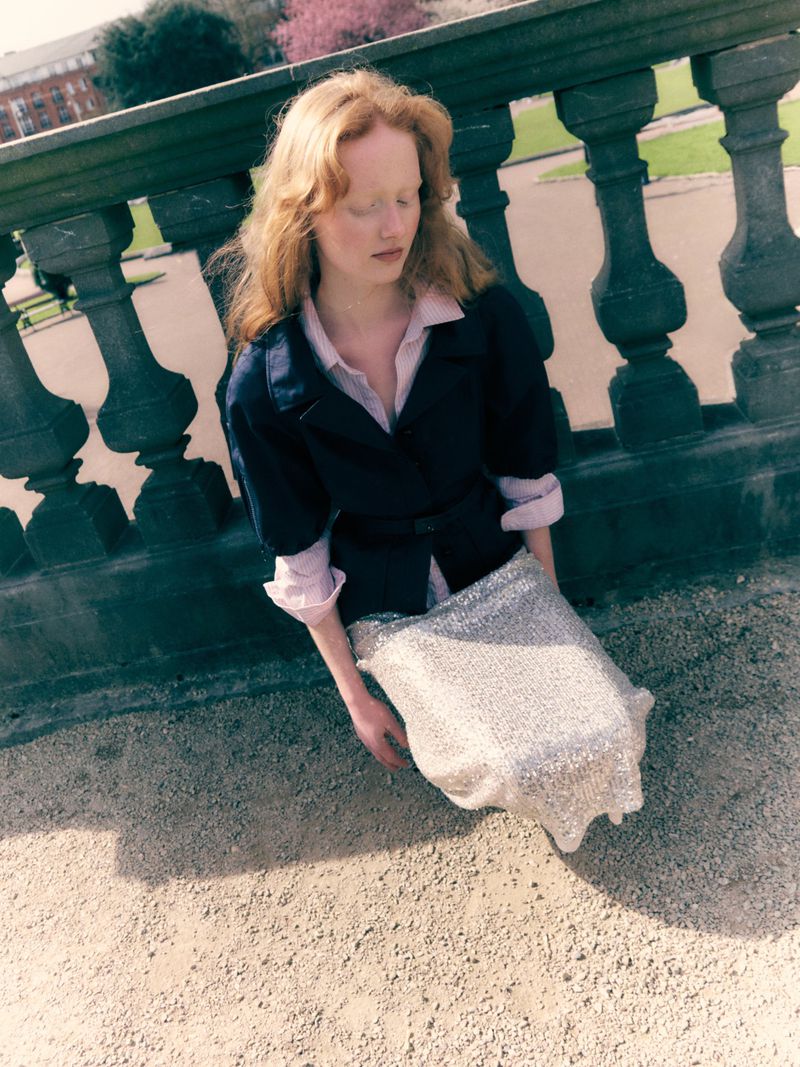 Model Lucy Arbuthnott wearing a black belted jacket and sparkling silver sequin skirt leaning against a classical stone balustrade.