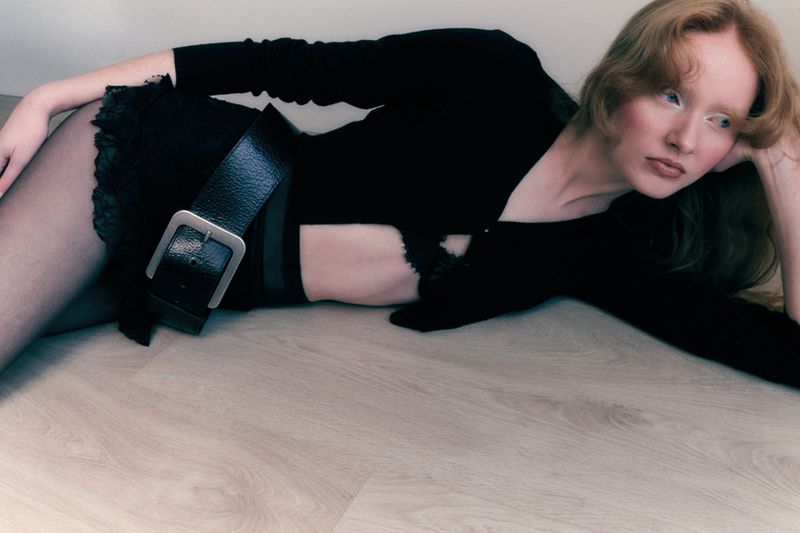 Model Lucy Arbuthnott reclining on a light wood floor wearing a black crop cardigan, lace mini skirt, and wide buckled leather belt.