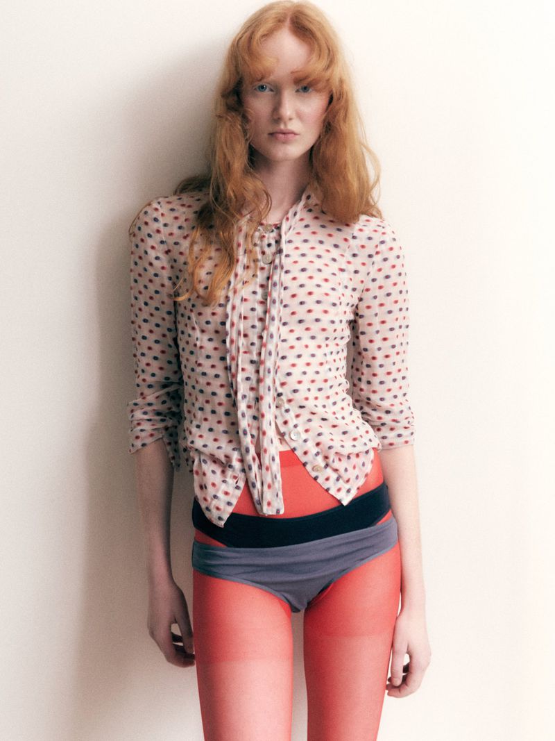 Model Lucy Arbuthnott posing in a patterned button-down shirt, layered briefs, and bright red tights against a white backdrop.