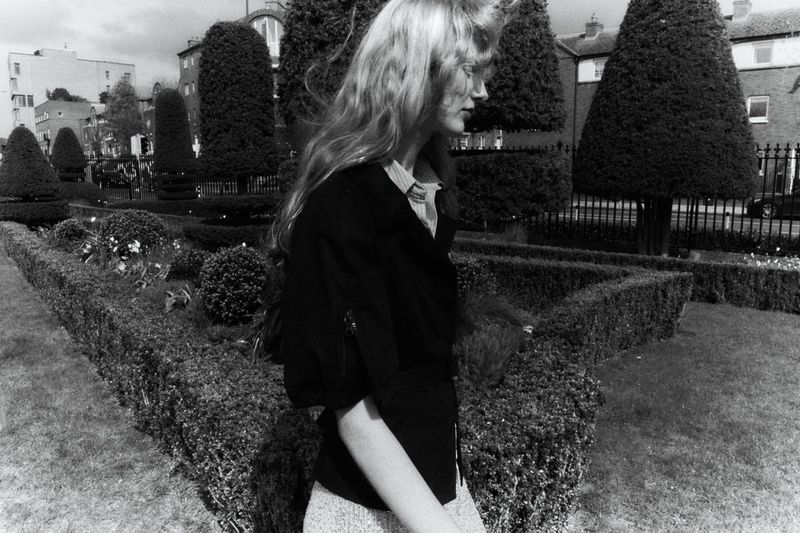 Black and white editorial portrait of model Lucy Arbuthnott walking past landscaped topiary bushes and residential buildings.
