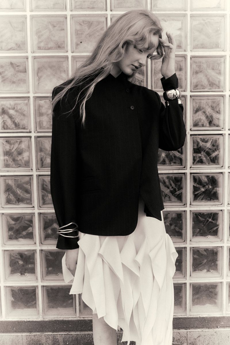 Lucy Arbuthnott posing in a black buttoned jacket and layered white handkerchief skirt against a glass block wall.