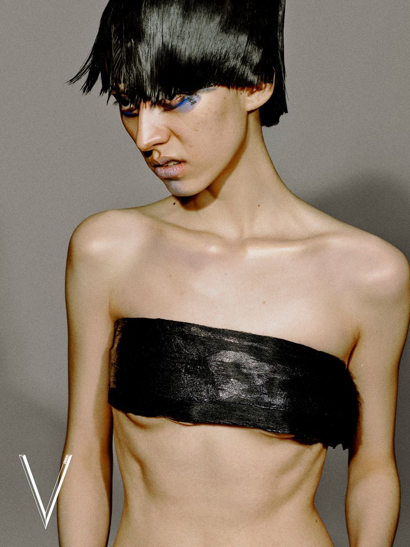 Model with chopped black fringe and blue eye makeup wearing a black strapless chest band against a grey studio backdrop.