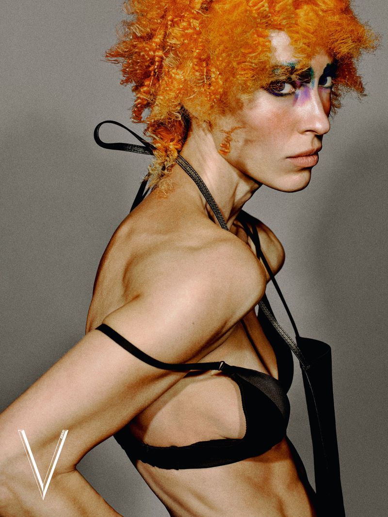 Model with bright orange hair and dramatic makeup wearing a black top against a grey studio backdrop