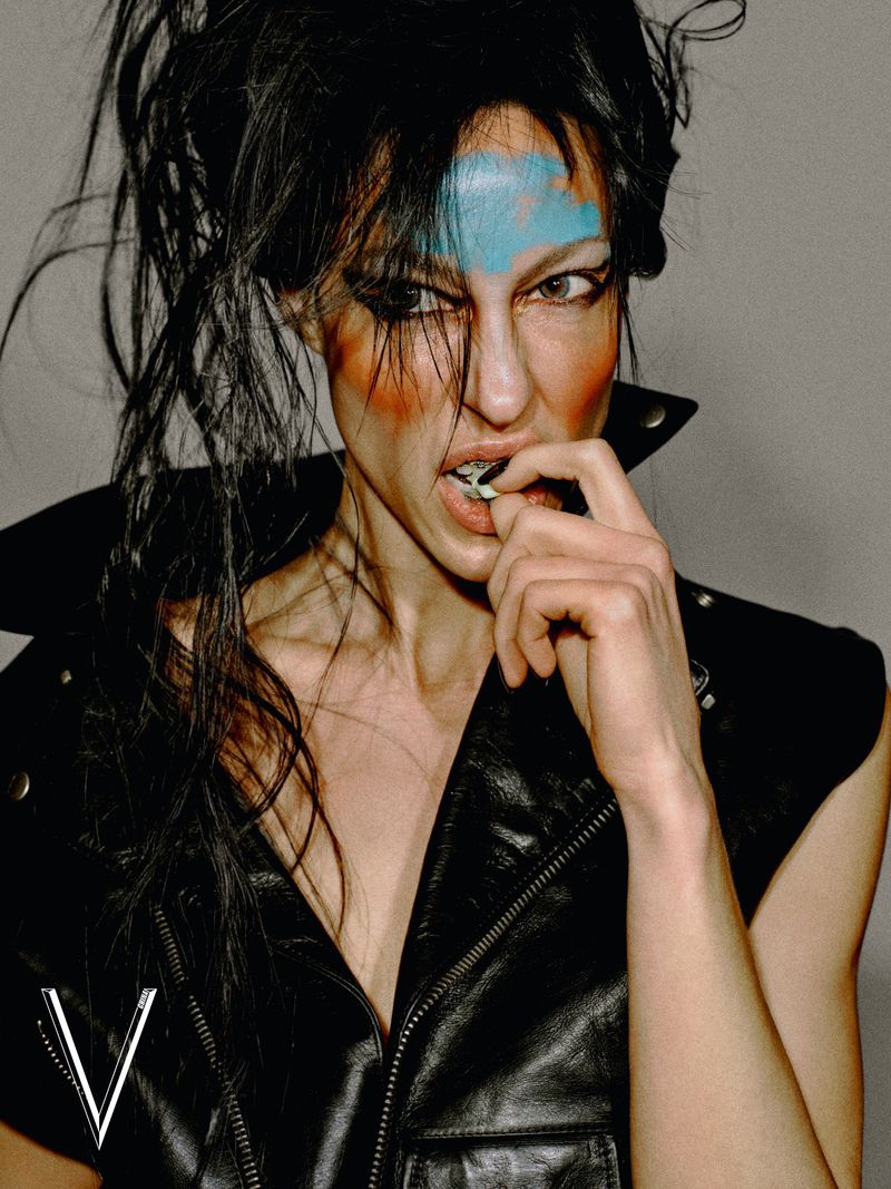 Model with avant-garde blue and orange makeup wearing a sleeveless black leather biker vest against a neutral studio backdrop.