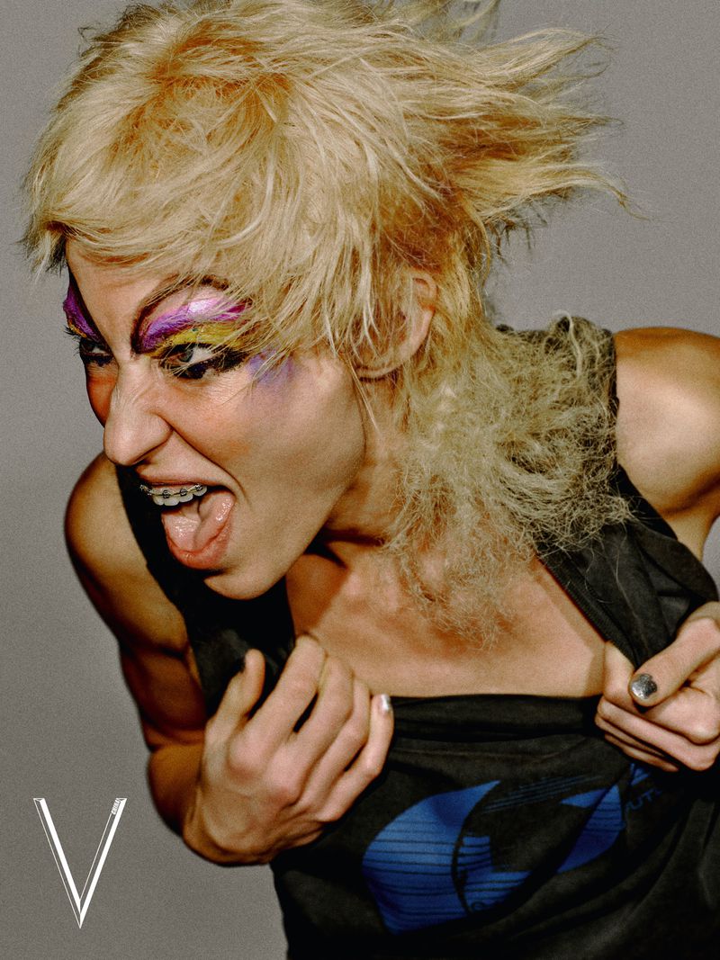 Model with blonde spiky hair and colorful eye makeup shouting while gripping a black graphic top against a grey background.
