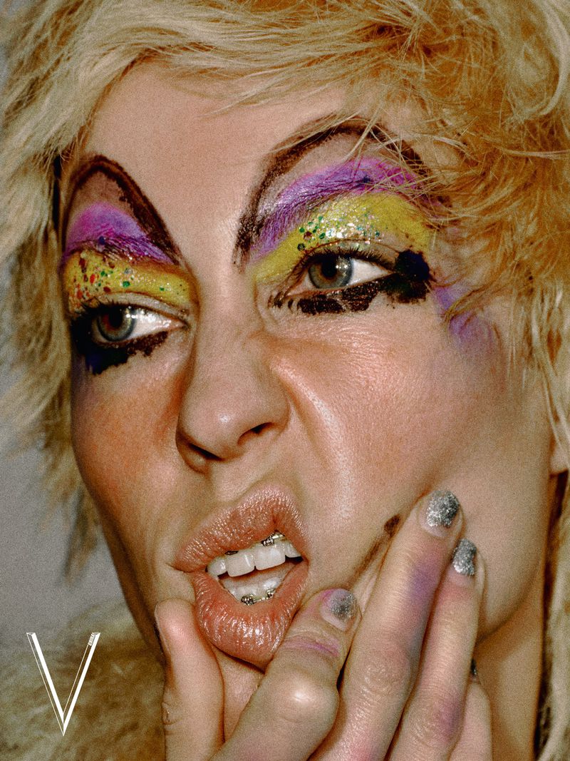 Close-up portrait of a blonde model with expressive theatrical eye makeup and silver glitter nails touching their cheek.