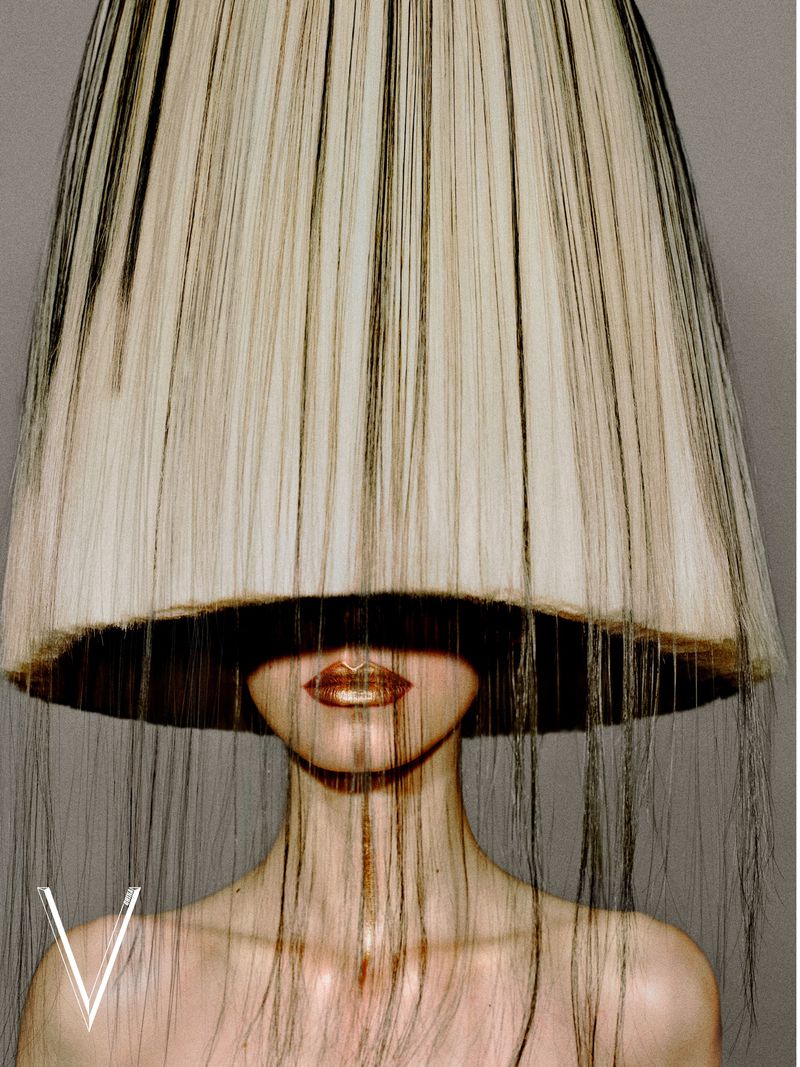 Model wearing a sculptural bell-shaped hairpiece covering the eyes, with metallic gold lips against a grey background.