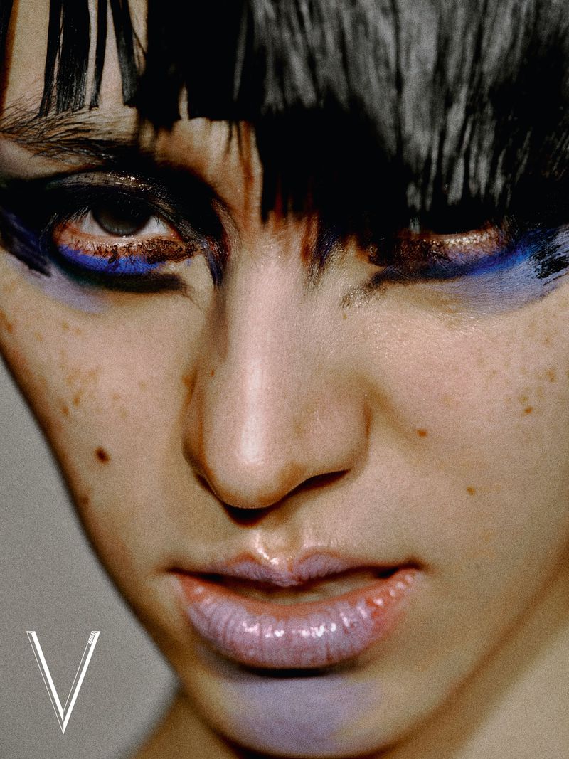 Extreme close-up beauty portrait of a model with dark fringe, bold blue and black eye makeup, and glossy lips.