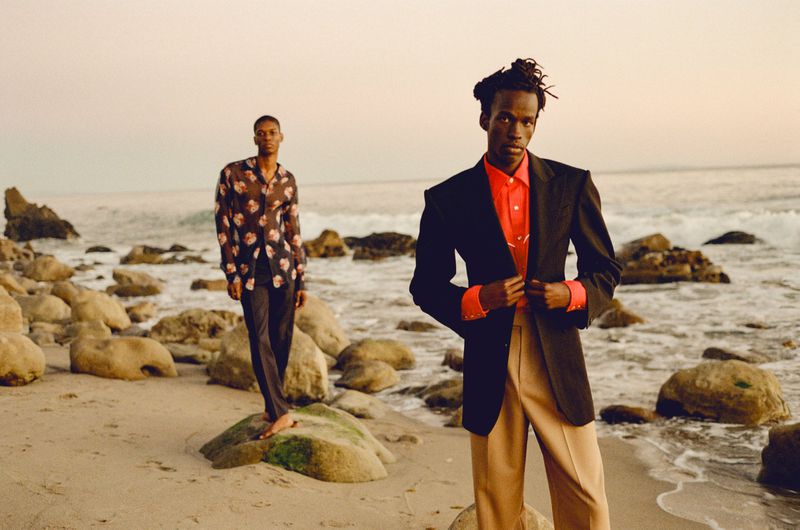 Two men posing on a rocky beach wearing tailored menswear, featuring a black blazer, red shirt, and beige trousers.