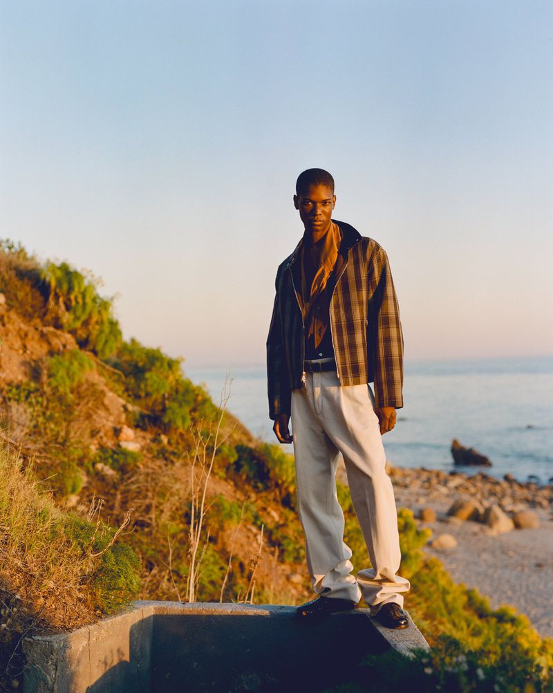 Model Kamari Moore standing on a concrete ledge in a plaid jacket and cream trousers along a rocky coast at sunset.
