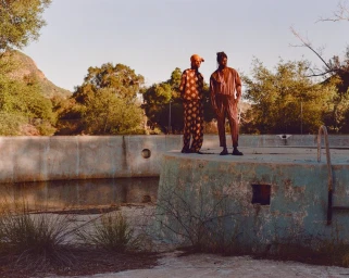 Two fashion models standing on the concrete edge of an abandoned outdoor swimming pool surrounded by trees.