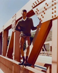 Model Kamari Moore in an olive green fringe sweater and black loafers posing on an orange steel truss bridge.