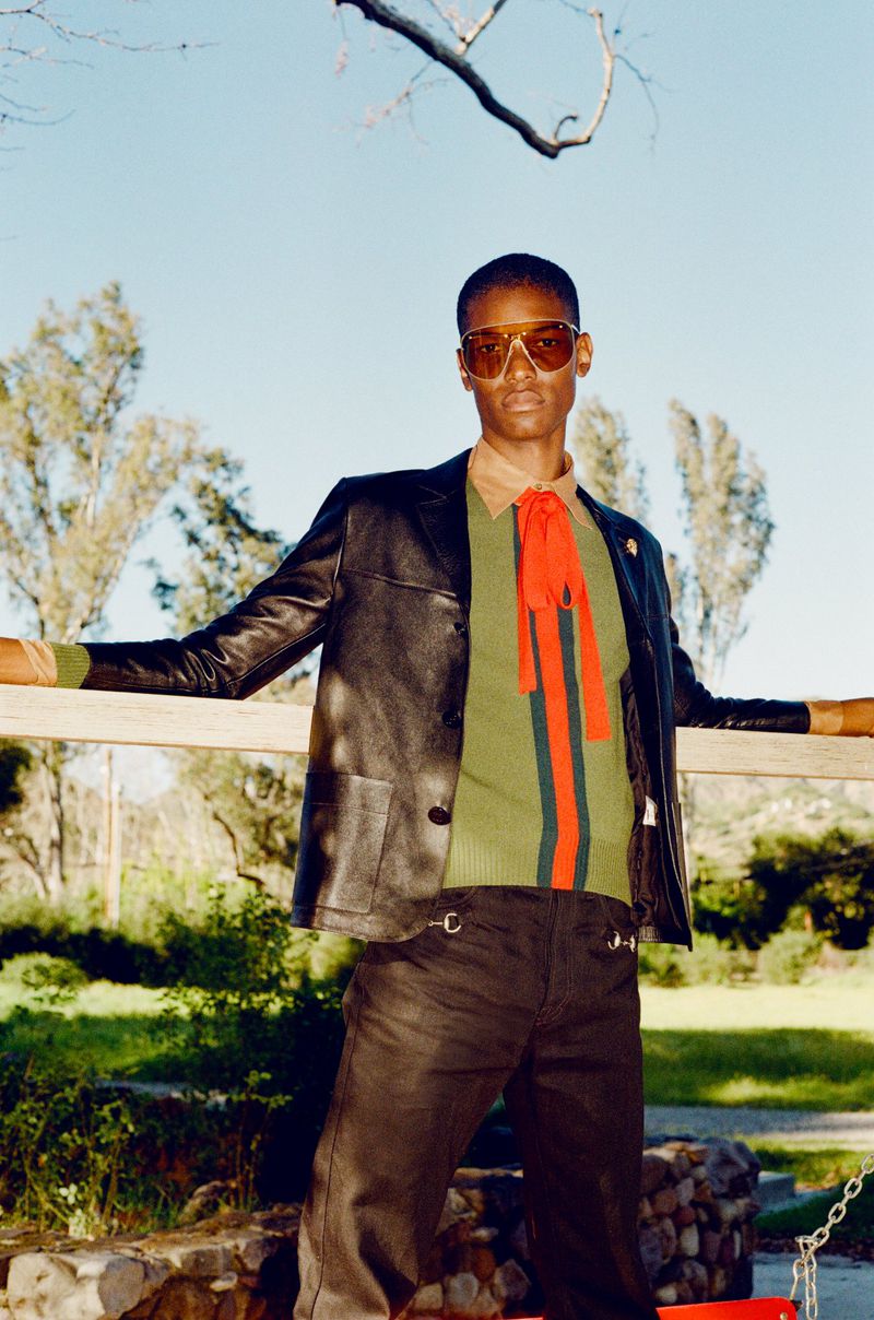 Model Kamari Moore wearing aviator sunglasses, a leather jacket, and an olive green knit top with a red tie against a wooden rail.