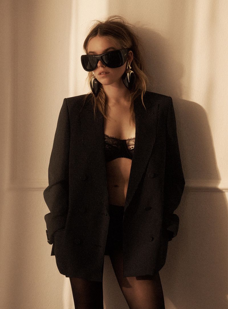 Actor Milly Alcock poses against a warm wall wearing an oversized black pinstripe blazer, lace bra, and large dark sunglasses.