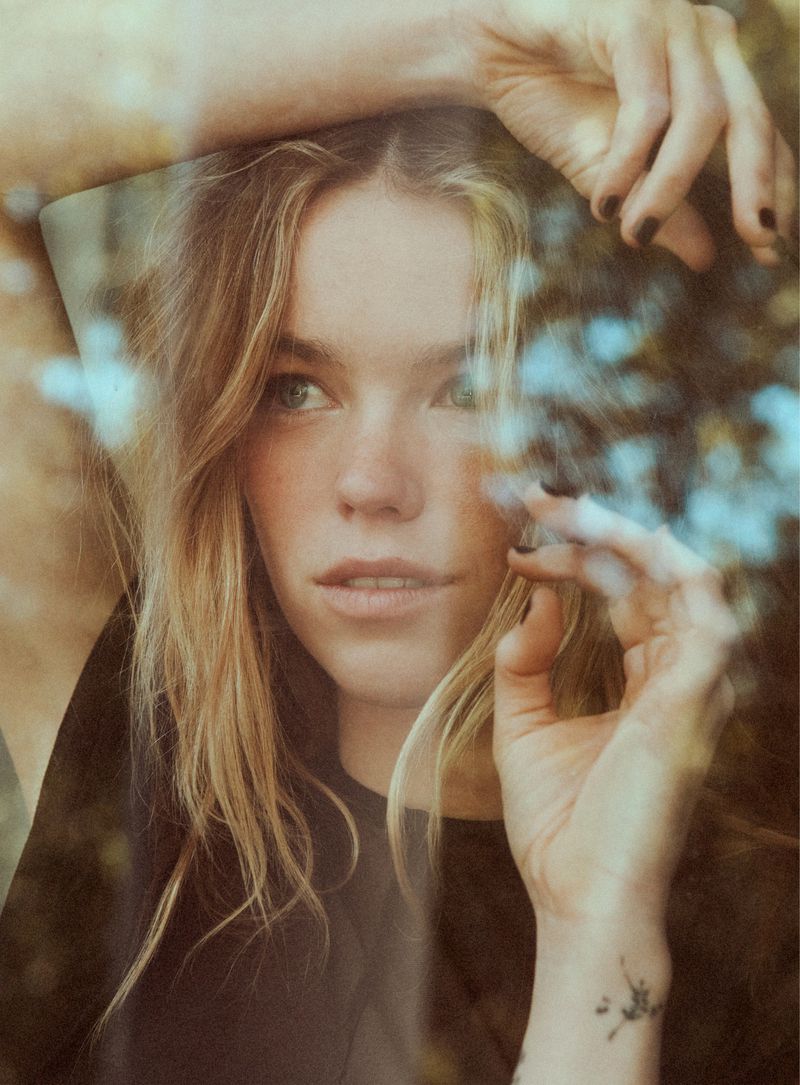 Close-up portrait of actor Milly Alcock resting her hands beside her face, seen through soft foreground reflections.