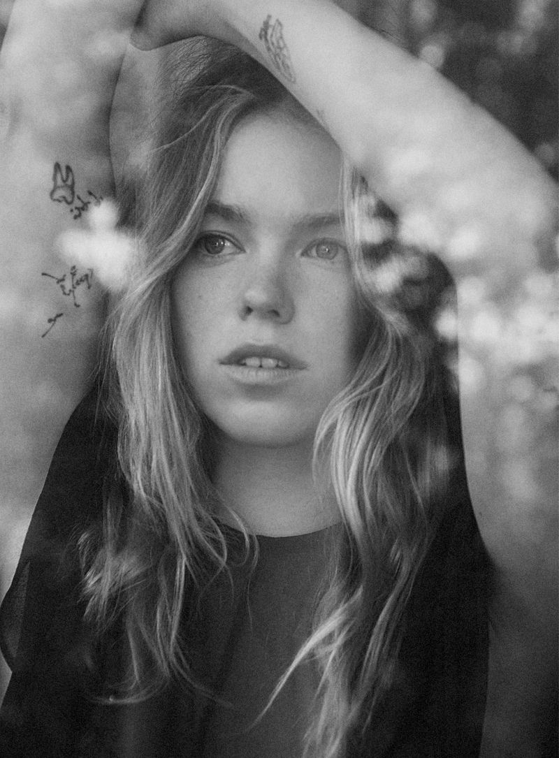 Monochrome close-up portrait of Milly Alcock with arms raised above her head behind blurred foreground reflections.