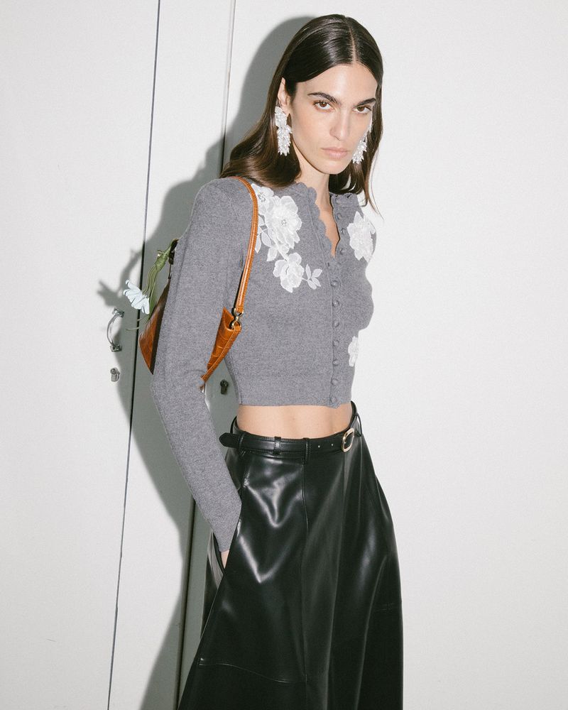 Model wearing a grey floral-applique cropped cardigan and black leather skirt standing against a white panelled wall.