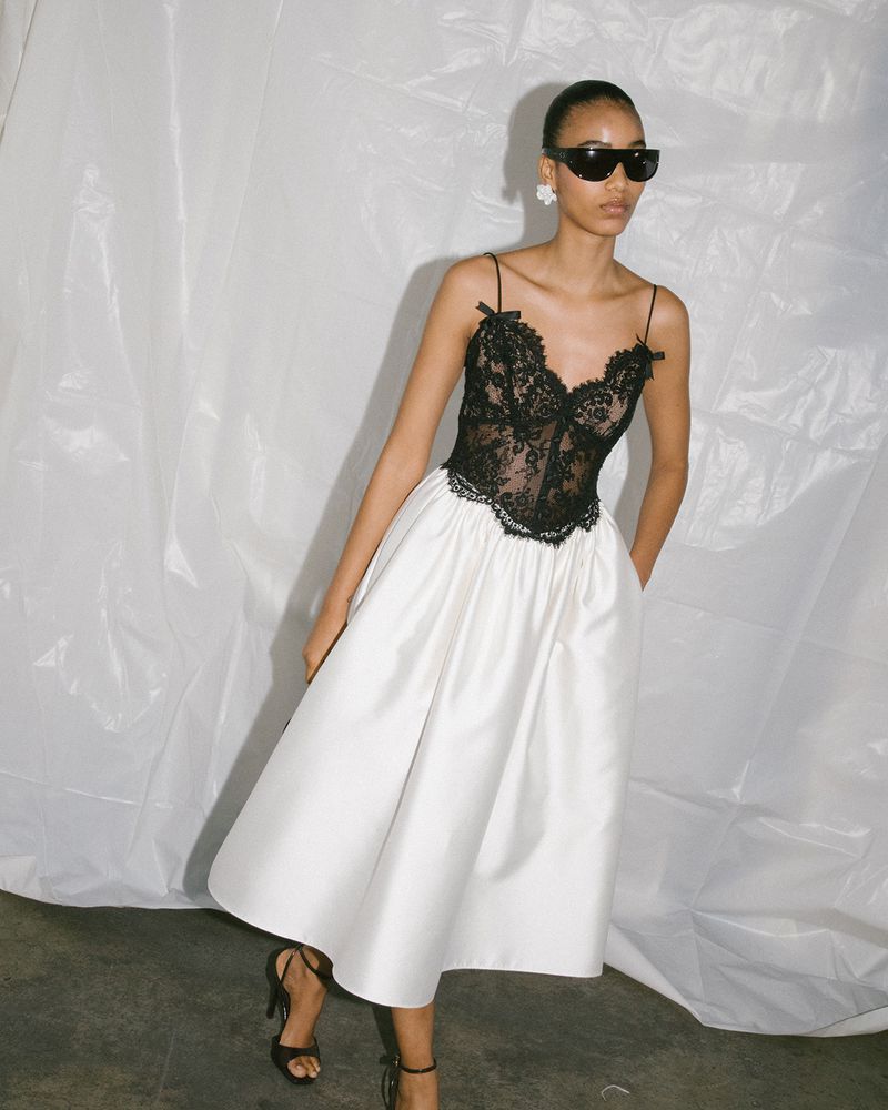 Fashion model in a black lace corset bodice and white satin midi skirt with wrap sunglasses against a white textured backdrop.
