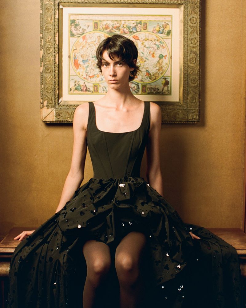 Model with short dark hair in a black corset dress with an embellished voluminous skirt, seated against a warm wall with a framed map.