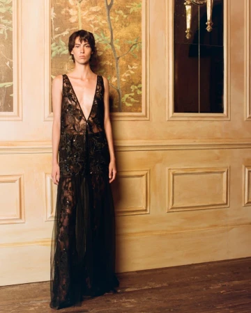 Model wearing a sheer embellished black plunging gown standing against paneled walls with floral art.