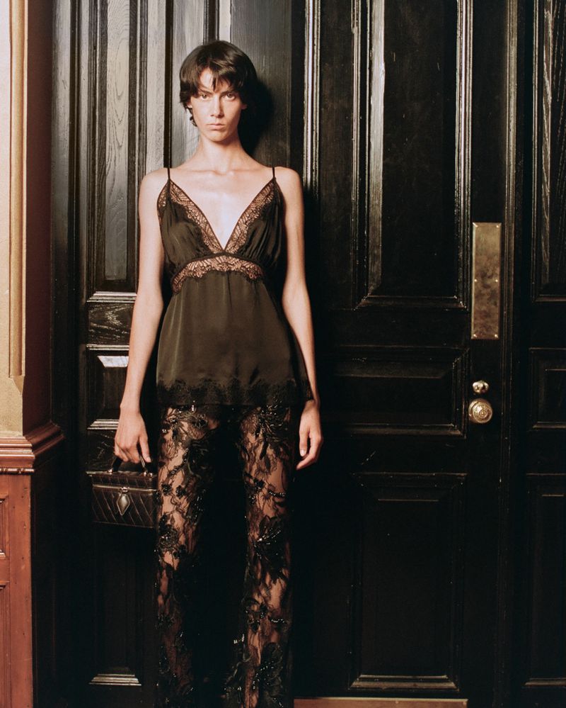 Model wearing a black lace-trimmed camisole and sheer black lace trousers holding a small box bag in front of a dark paneled door.