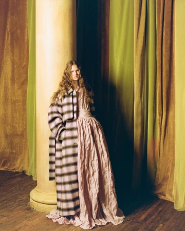 Model standing next to a large column wearing a long plaid coat and pink gown against draped curtains
