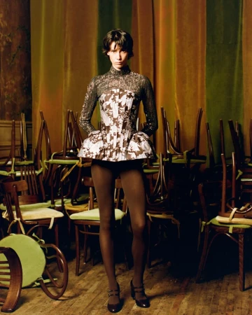 Model wearing a structured floral peplum top and brown tights standing among stacked wooden chairs against striped drapes.