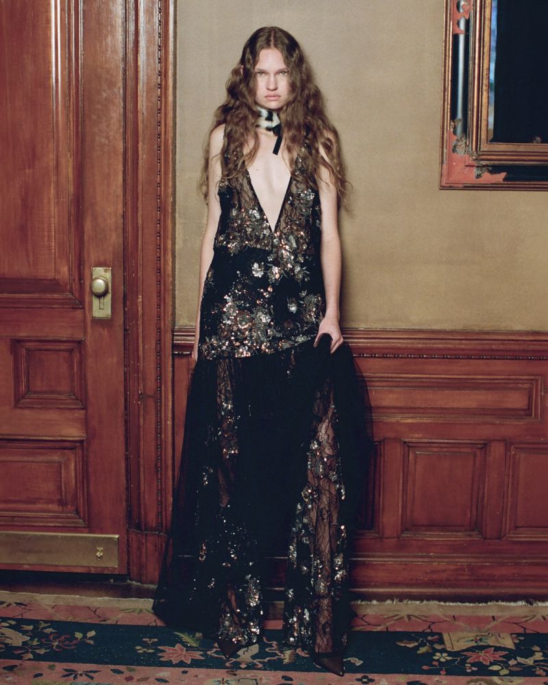 Model wearing a plunging black sequined evening gown with sheer lace against classic wood-paneled walls on a patterned carpet.