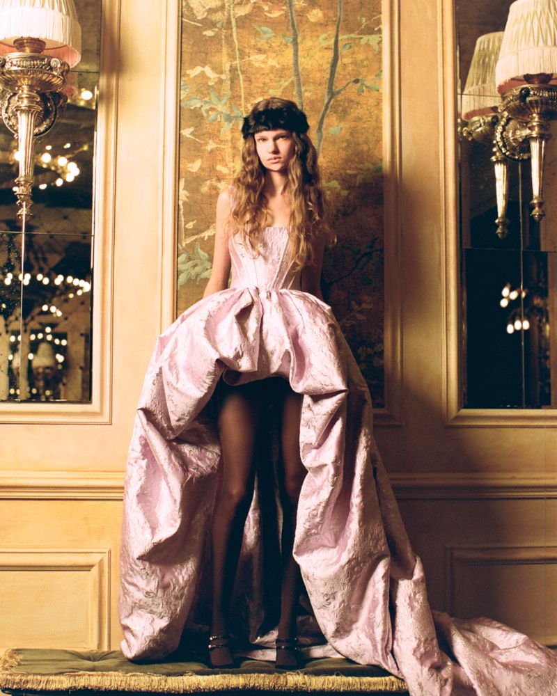Model wearing a strapless pale pink high-low ballgown with a long train and black tights, standing before an ornate paneled wall.