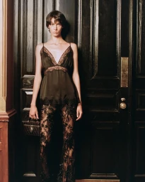 Model wearing a black lace-trimmed camisole and sheer black lace trousers holding a small box bag in front of a dark paneled door.