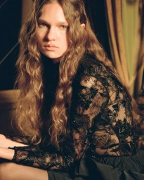 Model with long wavy brown hair wearing an embroidered black sheer lace top seated in a dimly lit room with drapery.