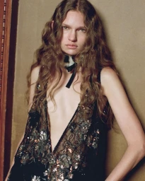 Model with long wavy hair wearing a plunging sequin-embroidered black lace garment and a fur choker necklace against a wall.