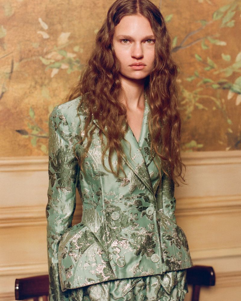 Model with long wavy brown hair wearing a mint green metallic floral brocade tailored jacket against a painted wall.