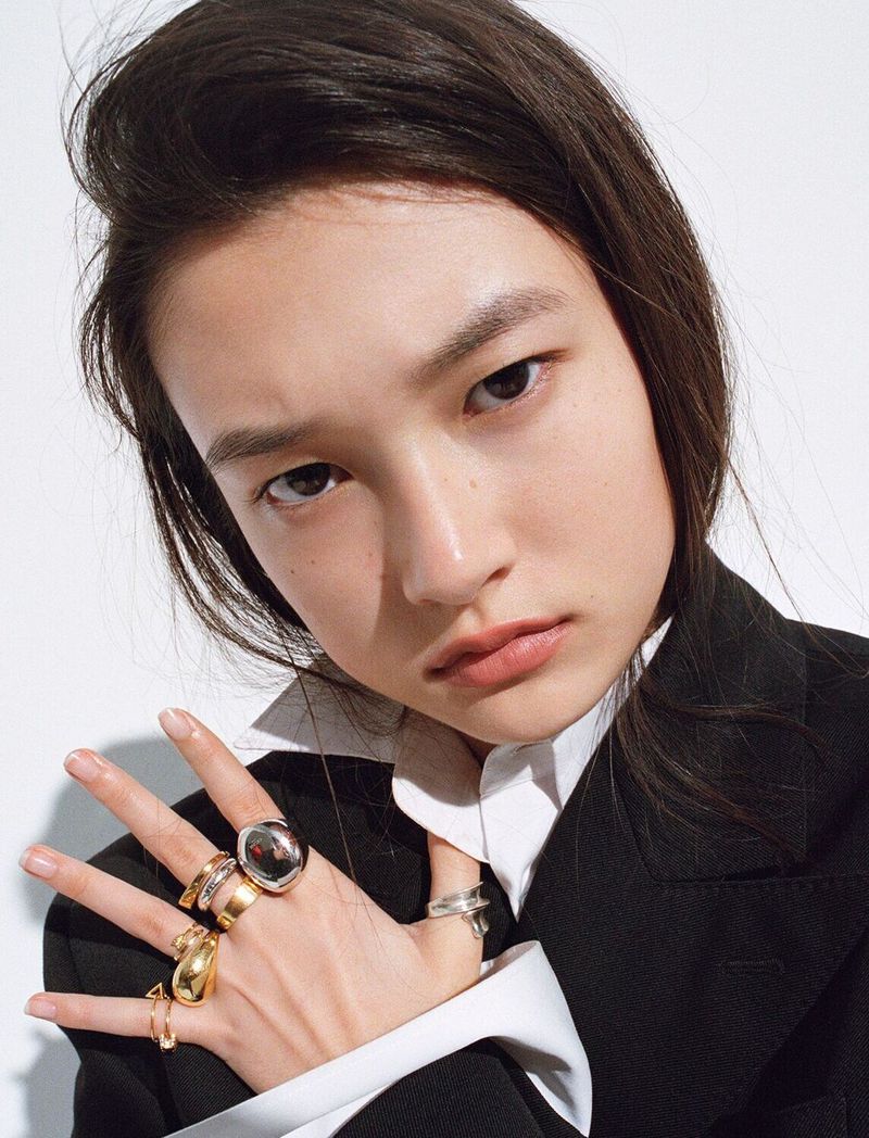 Model Carolyn Pan wearing a black blazer, white collared shirt, and multiple sculptural rings against a white background.