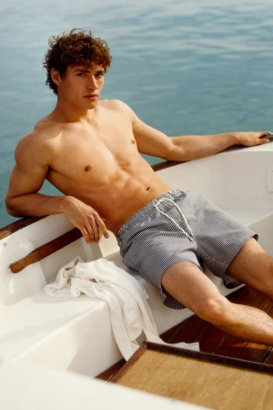 Valentin Humbroich reclining shirtless in patterned swim shorts on a boat on open water.