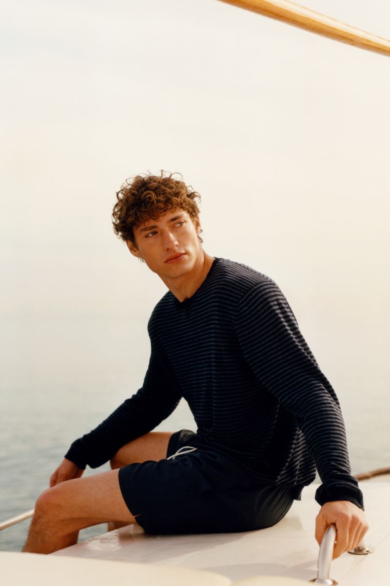Valentin Humbroich in a dark long-sleeve striped knit and swim shorts seated on a boat deck overlooking calm open water.