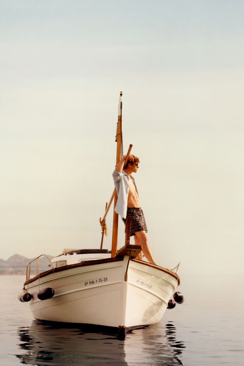 Model Valentin Humbroich wearing swim shorts and an unbuttoned shirt while standing on the bow of a boat on calm water.