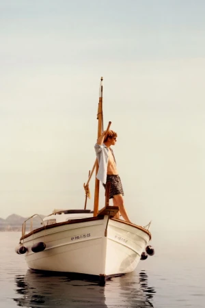 Model Valentin Humbroich wearing swim shorts and an unbuttoned shirt while standing on the bow of a boat on calm water.