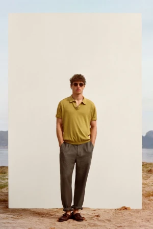 Man wearing a green polo shirt and grey trousers standing in front of a white panel on a sandy beach
