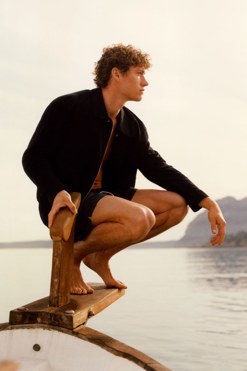 Model Valentin Humbroich crouching barefoot on a wooden boat bow over calm water in a black jacket and shorts.