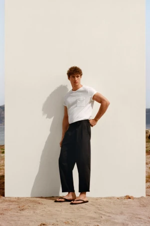 Model Valentin Humbroich wearing a white pocket t-shirt and black cropped trousers against a white backdrop panel on sand.