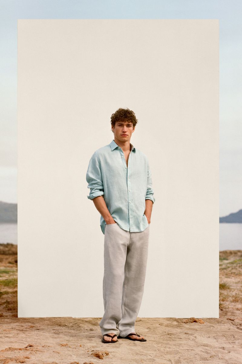 Valentin Humbroich posing in a light blue linen shirt and relaxed trousers before a white panel on a sandy shore.