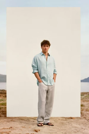 Valentin Humbroich posing in a light blue linen shirt and relaxed trousers before a white panel on a sandy shore.