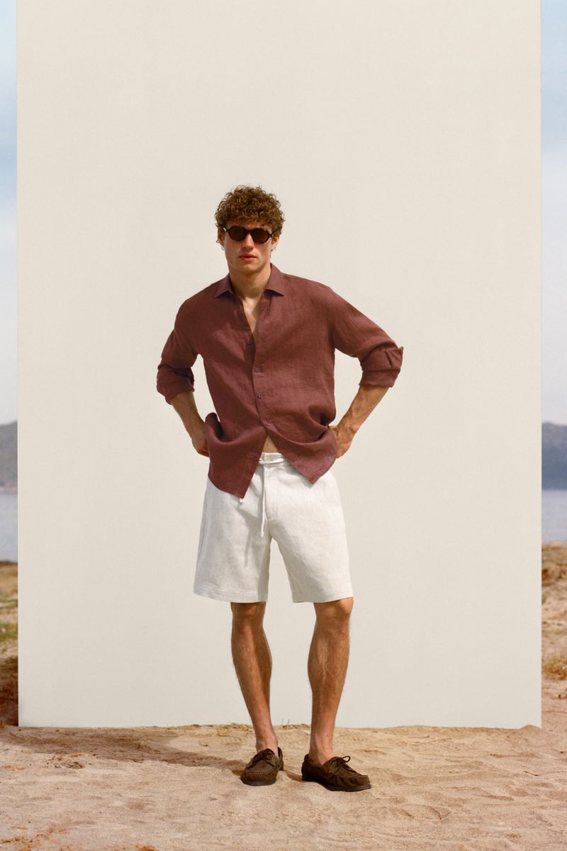 Model Valentin Humbroich in a terracotta linen shirt, white shorts, and sunglasses standing in front of a white monolith on sand.