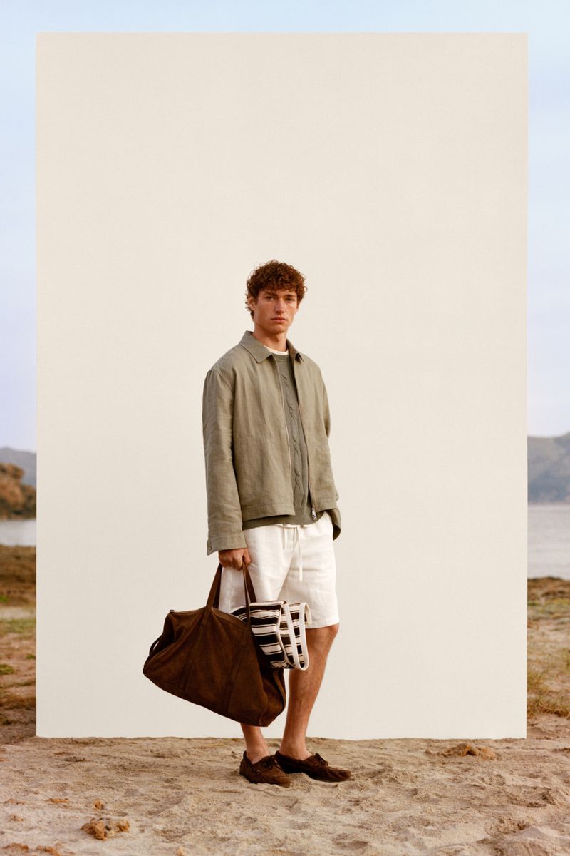 Male model wearing green jacket and white shorts holding a dark brown bag standing outdoors against a white panel on sand.